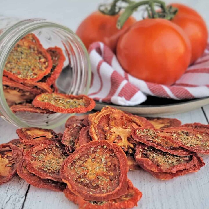 Seasoned dried tomato chips spilling from a mason jar, fresh vine ripe tomatoes on red striped towel in background.