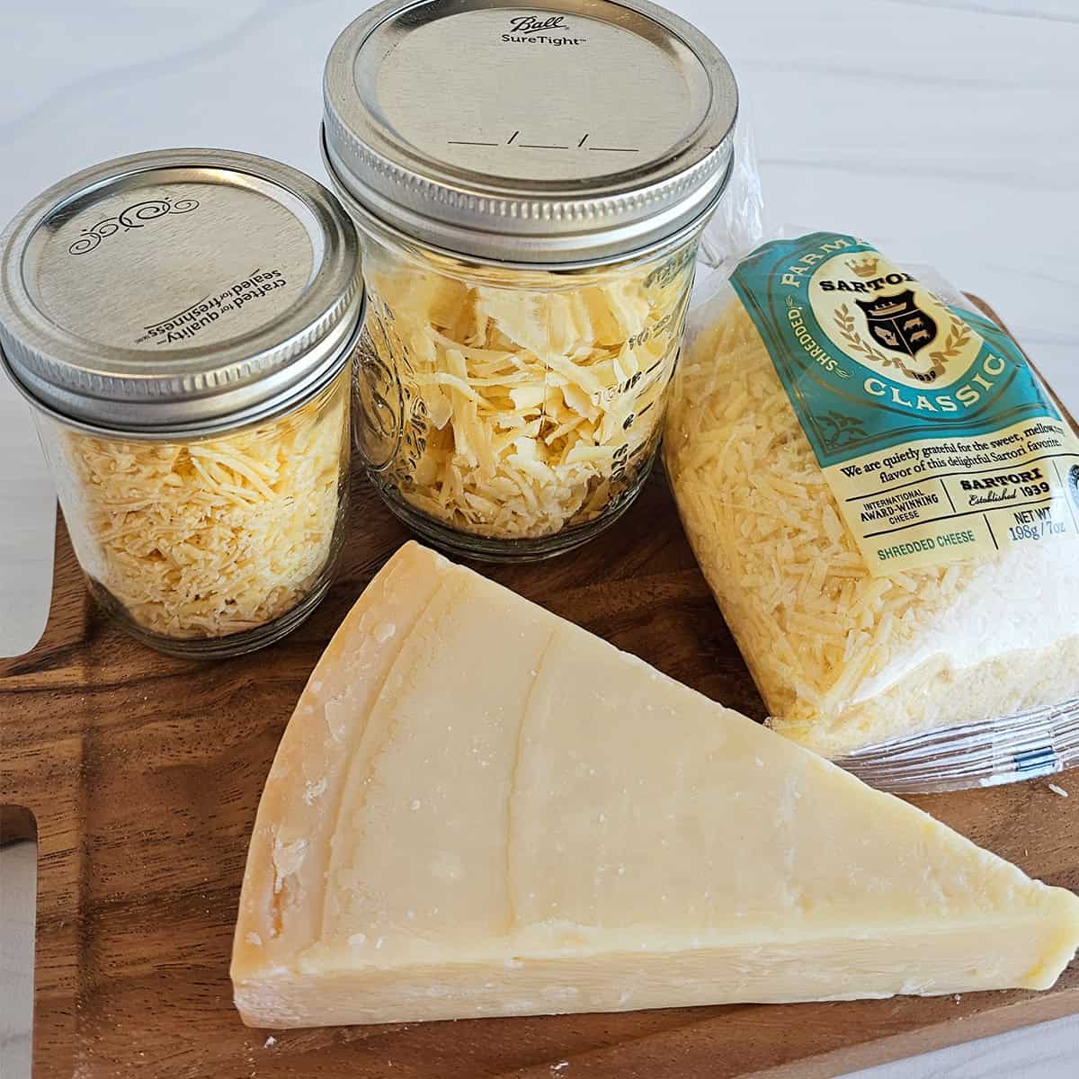 A wood cutting board with a wedge of Parmesan cheese, two jars of dehydrated Parmesan, and a bag of shredded Parmesan cheese.