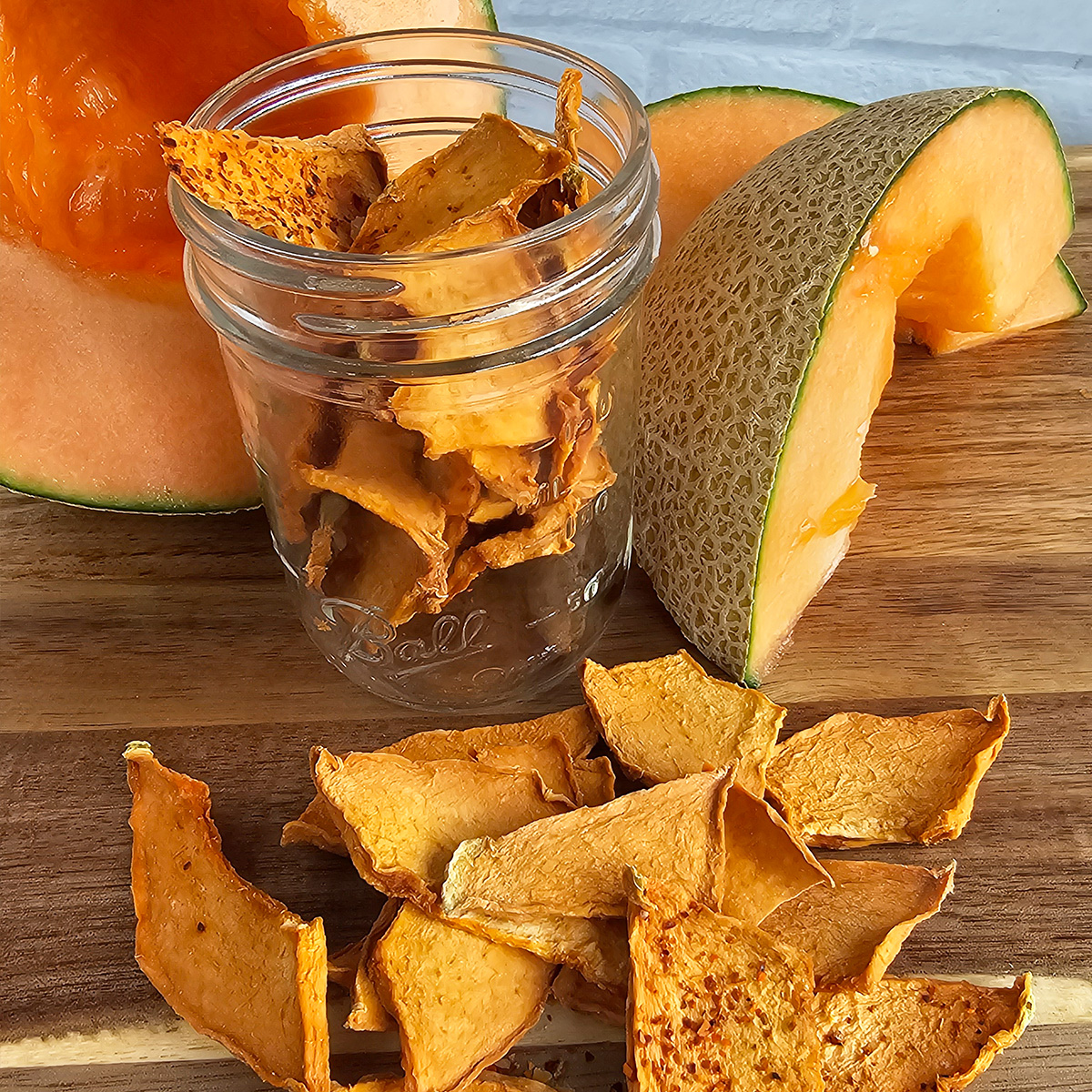 Dried cantaloupe pieces scattered on a wooden surface, with a glass jar filled with more dried cantaloupe pieces, and fresh cantaloupe slices in the background.