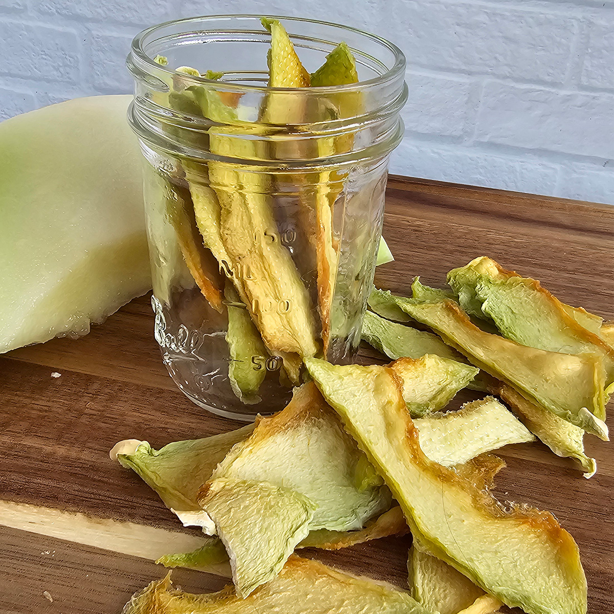 A jar of dried honeydew melon with scatter pieces on a wooden cutting board and a slice of fresh in the background.