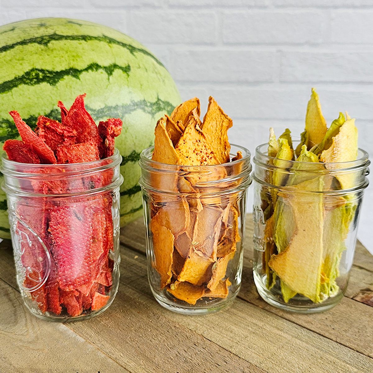 Three glass jars filled with dehydrated watermelon, cantaloupe and honeydew slices placed on a wooden surface. A whole watermelon is positioned behind the jars.